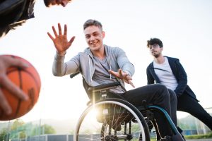 A boy in wheelchair with teenager friends outside playing basketball.