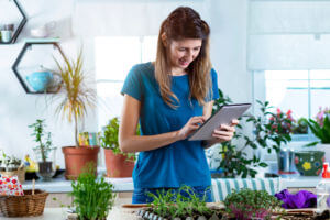 A woman in a florist shop surrounded by plants and flowers, reading from a tablet.