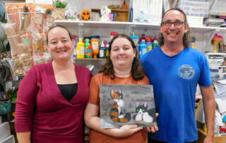 Emily proudly displays her cat painting with her parents. Bonnie, Emily's mother, beams with joy at her daughter's creative accomplishments in visual art.