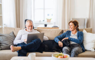 Man and woman sitting on couch with their dog between them. Man on computer, woman smiling at man.