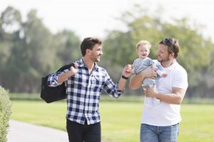 Two men walking in a park together, one holding a baby and the other engaging with the baby